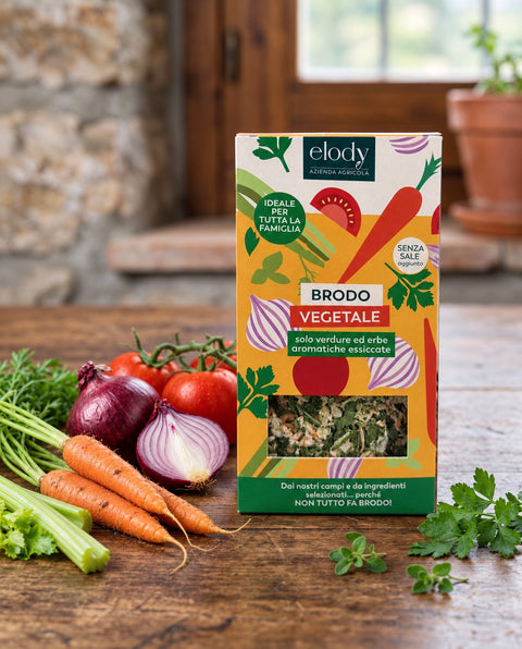 Confezione di Brodo vegetale Elody sul tavolo di cucina con carote, cipolle rosse, pomodori e sedano